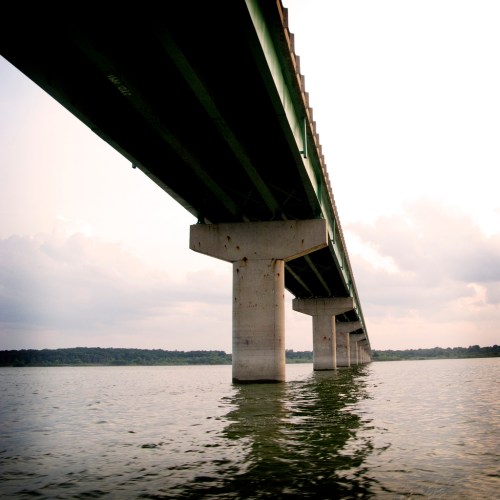 A low-angle view of a bridge spanning over calm water, with a cloudy sky in the background.