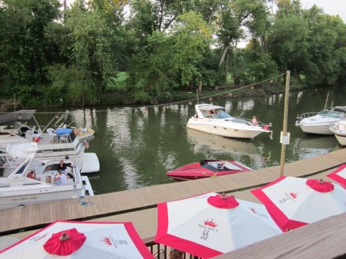 A view of a calm river with boats and umbrellas on a wooden dock, surrounded by lush green trees.