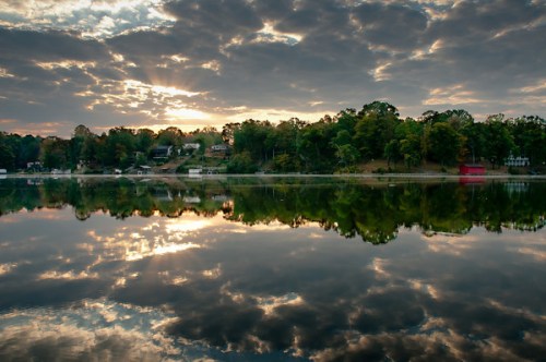 A serene lake at sunrise, reflecting clouds and trees, with a peaceful shoreline and colorful sky.