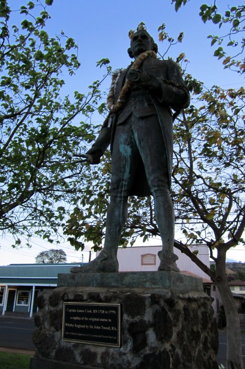 Statue of Captain James Cook, with a plaque, surrounded by trees and buildings in the background.