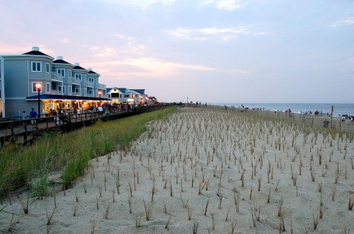 A beachside scene at dusk, featuring sandy dunes, grass, and a boardwalk lined with buildings and people.