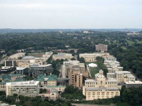Aerial view of a university campus surrounded by greenery and buildings, with a distant city skyline.