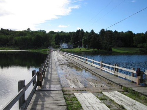 A wooden dock extends over calm water, leading to a grassy area with trees and a house in the background.