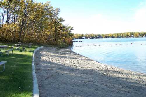 A sandy beach by a calm lake, surrounded by trees with autumn foliage and picnic tables nearby.