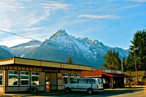 A small town scene featuring a building with large windows, a white van, and snow-capped mountains in the background.
