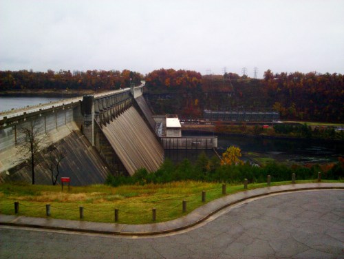 A concrete dam with a sloped face, surrounded by trees with autumn foliage and a river below, under a cloudy sky.