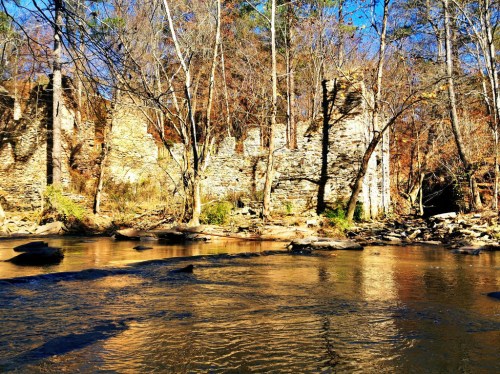 Ruins of a stone structure beside a calm river, surrounded by trees and autumn foliage under a clear blue sky.