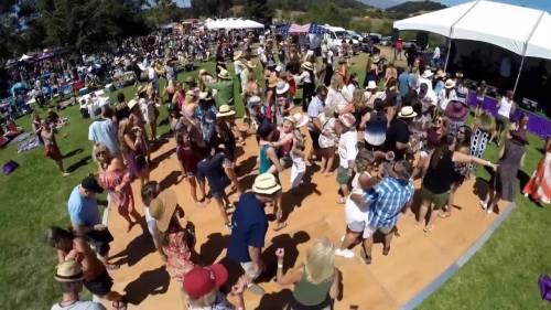 A lively outdoor festival scene with a large crowd dancing on a wooden floor, surrounded by greenery and tents.
