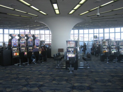 A spacious airport area with slot machines and large windows, showing travelers and a modern design.