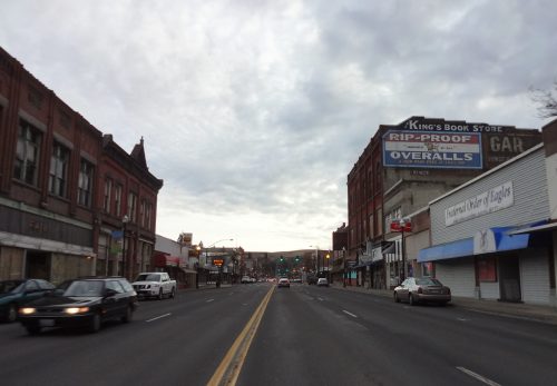 A street view featuring shops, cars, and a cloudy sky, with signs for a bookstore and overalls store.