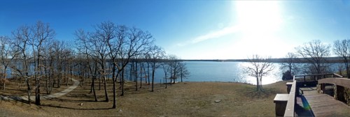 Panoramic view of a serene lake surrounded by bare trees under a clear blue sky. Sunlight reflects on the water.