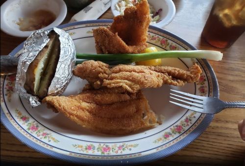 A plate with fried fish, a baked potato wrapped in foil, a green onion, and a lemon wedge.
