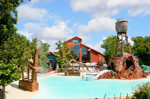 A water park scene featuring a large pool, water slides, and a rustic building in the background under a blue sky.