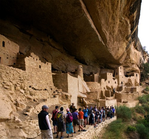 A group of people stands in front of ancient cliff dwellings, surrounded by rocky terrain and greenery.