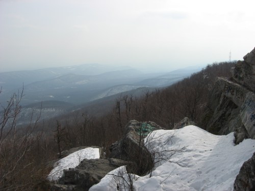 A scenic view from a rocky overlook, featuring distant mountains and patches of snow on the ground.