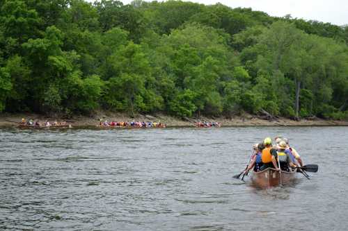 A group of people paddling a canoe on a river, surrounded by lush green trees and another canoe group in the background.