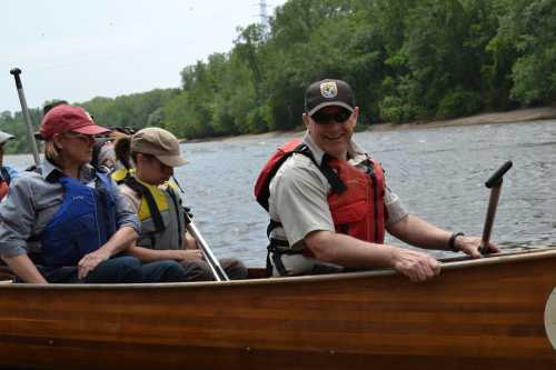 A group of people in life jackets smiles while paddling a canoe on a river surrounded by greenery.