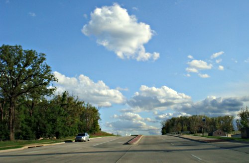 A wide road stretches ahead under a blue sky filled with fluffy clouds, flanked by trees on one side.