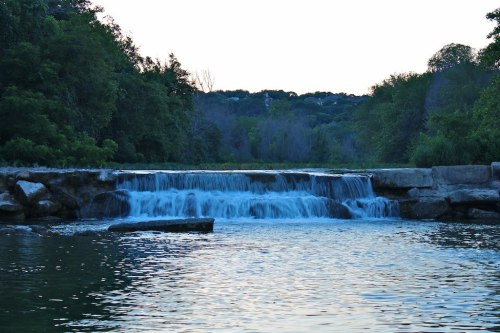 A serene waterfall cascades over rocks into a calm river, surrounded by lush greenery and trees.