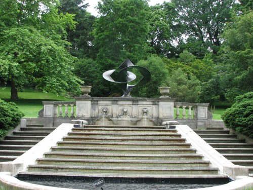 A modern sculpture atop a stone fountain, surrounded by lush greenery and steps leading down to a water feature.
