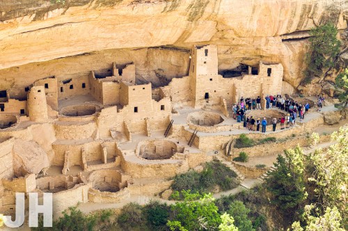 An ancient cliff dwelling with stone structures, surrounded by greenery and a group of visitors exploring the site.