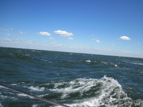 Waves on a blue ocean under a clear sky with scattered clouds, viewed from a boat.