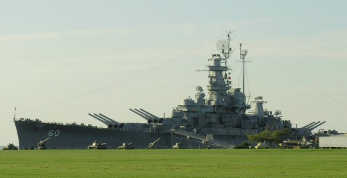 A large battleship with multiple gun turrets, docked near a grassy area under a clear sky.