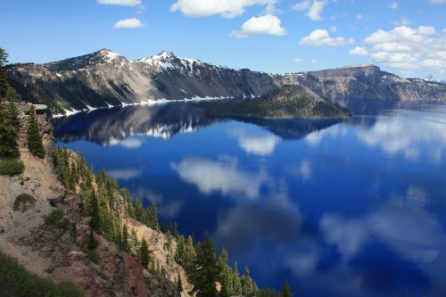 A serene view of Crater Lake, with blue waters reflecting clouds and mountains under a clear sky. Pine trees in the foreground.
