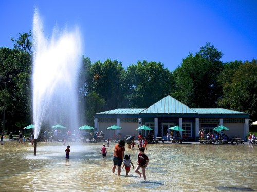 Children play in a splash pad with a fountain, while adults relax under umbrellas near a building in a park.