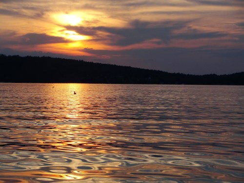 A serene sunset over a calm lake, with rippling water reflecting warm hues and a silhouette of distant hills.