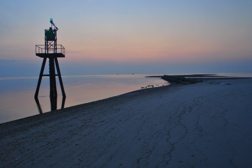 A lighthouse stands on stilts by a calm sea at dusk, reflecting soft pastel colors in the water.