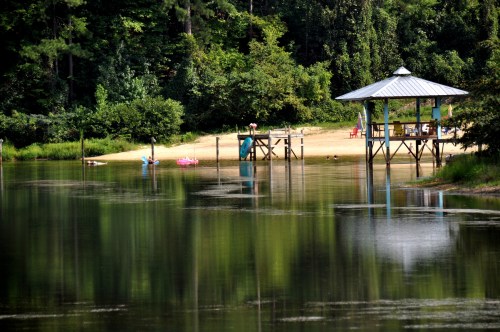 A serene lake scene with a gazebo, sandy beach, and lush greenery reflecting in the calm water.
