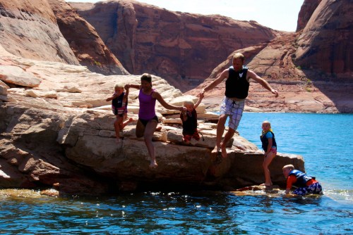 A family jumps off a rock into a blue lake, surrounded by red rock formations on a sunny day.