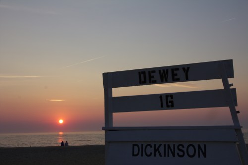 Lifeguard stand at sunset on a beach, with the sun setting over the water and silhouettes of people in the distance.