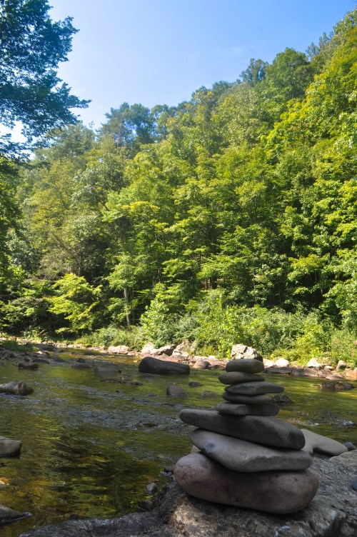 A stack of smooth stones by a serene river, surrounded by lush green trees under a clear blue sky.