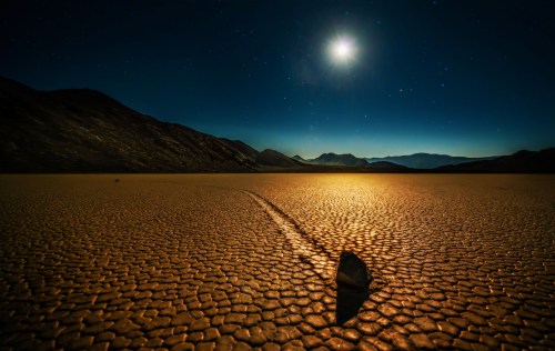 A moonlit desert landscape with a rock leaving a trail on cracked earth, surrounded by mountains and a starry sky.