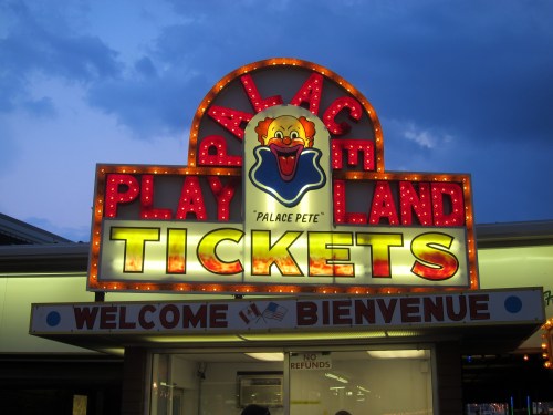 Brightly lit sign for "Palace Playland" featuring a clown, with "Tickets" and "Welcome Bienvenue" displayed.