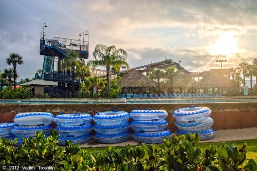 A water park scene featuring stacked blue inner tubes, palm trees, and a tall water slide in the background.