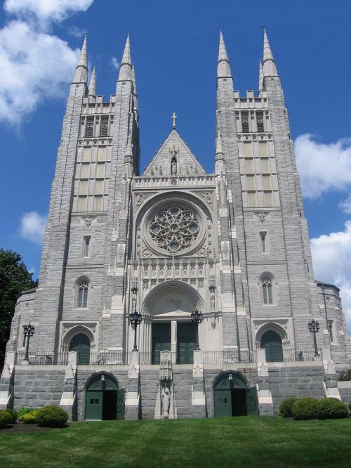 Gothic-style church with tall spires, intricate rose window, and green doors, set against a blue sky with clouds.