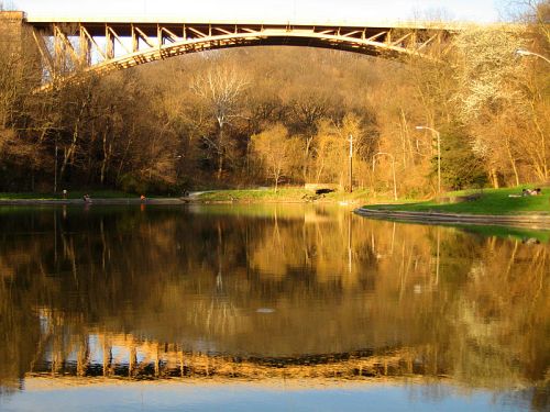 A bridge reflected in a calm river, surrounded by trees and greenery under a clear sky.