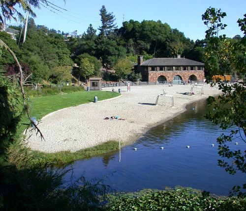 A sandy beach area beside a calm lake, with a historic building and trees in the background.