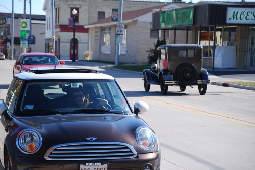 A modern car drives past a vintage car on a street lined with shops and buildings.
