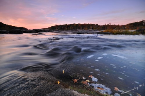 A serene river scene at sunset, with flowing water, rocks, and autumn leaves along the shore.
