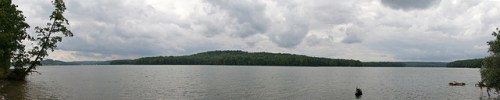 A panoramic view of a calm lake surrounded by lush green hills under a cloudy sky.