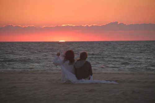 Two people sit on the beach, watching a vibrant sunset over the ocean, wrapped in a white blanket.