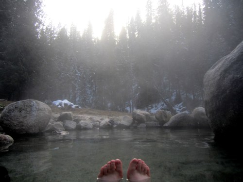 Feet submerged in a steaming hot spring, surrounded by misty trees and large rocks.
