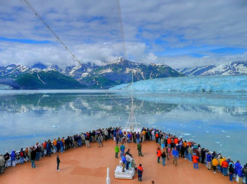 A crowd of people on a ship's deck, gazing at a glacier and mountains under a cloudy sky.