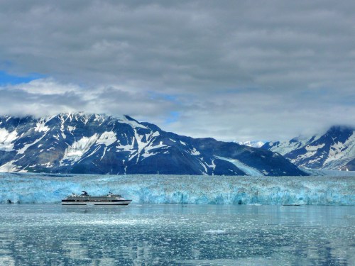 A cruise ship floats near a massive glacier, surrounded by snow-capped mountains and a cloudy sky.