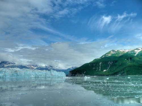 A serene landscape featuring a glacier, calm waters, and lush green mountains under a partly cloudy sky.