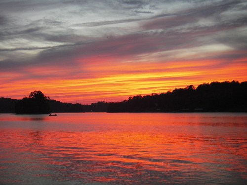 A vibrant sunset over a calm lake, with orange and pink hues reflecting on the water and silhouettes of trees in the background.